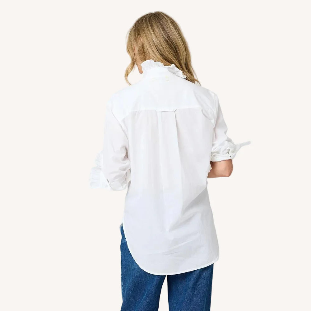 A person with long blonde hair faces away, wearing the Kerri Rosenthal Mia Ruffle Shirt—a white women's blouse with a ruffled collar and long sleeves—paired with blue jeans against a plain light background.