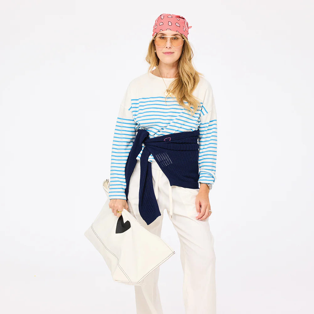 Wearing the Kerri Rosenthal Artist Long Sleeve Stripe Tee in blue and white, a woman pairs it with white pants, a navy sweater at her waist, glasses, and a large white bag with a black heart, all set against a plain white background.
