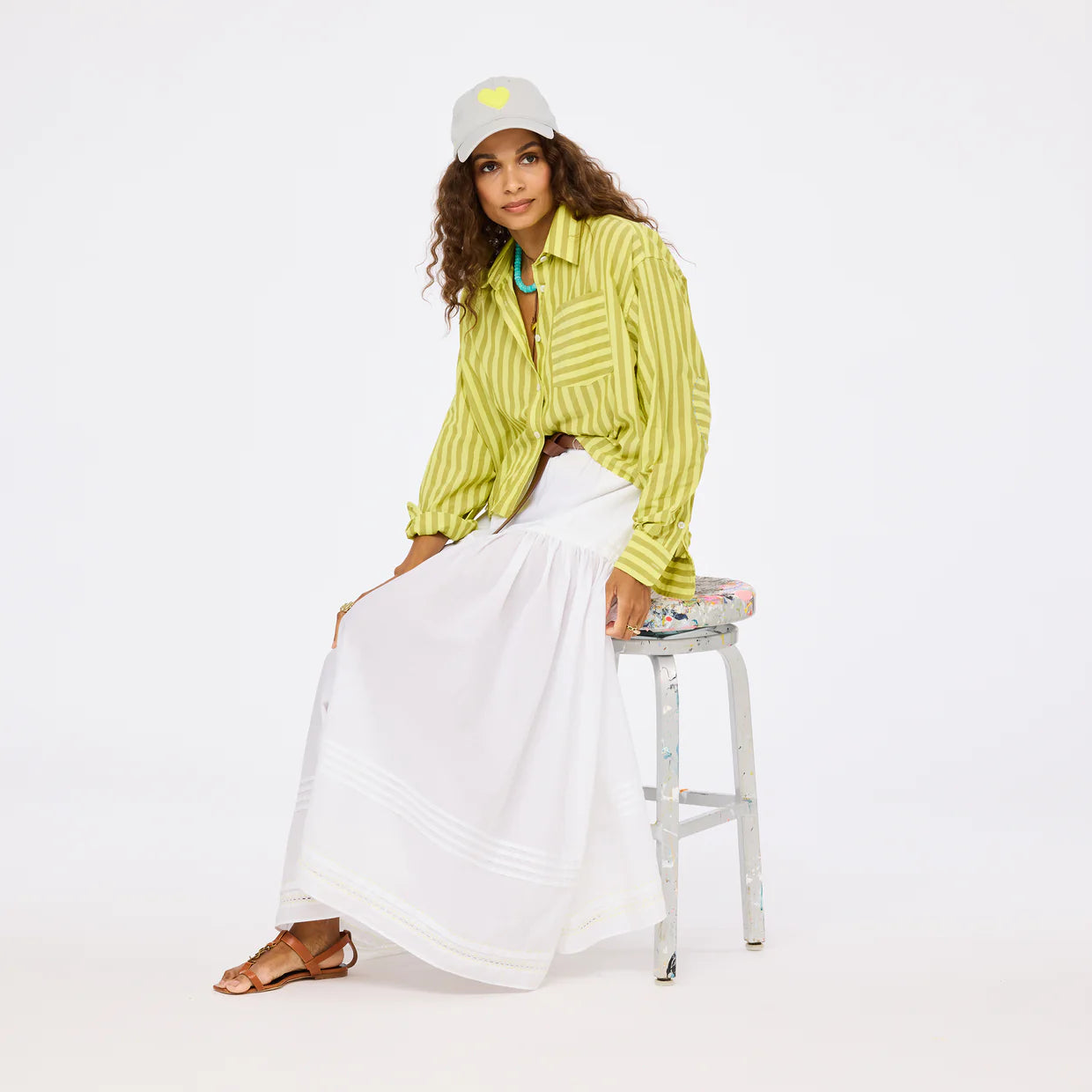 A woman sits on a stool in a Kerri Rosenthal Tia Faded Stripe Shirt with a long white skirt, brown sandals, turquoise necklace, and gray cap with yellow heart. The background is plain white.