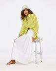 A woman sits on a stool in a Kerri Rosenthal Tia Faded Stripe Shirt with a long white skirt, brown sandals, turquoise necklace, and gray cap with yellow heart. The background is plain white.