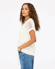 Woman with long curly hair wears the Kerri Rosenthal Summer Vestie Sweater, styled with blue jeans and gold jewelry, standing sideways with hands in her pockets against a plain white background.