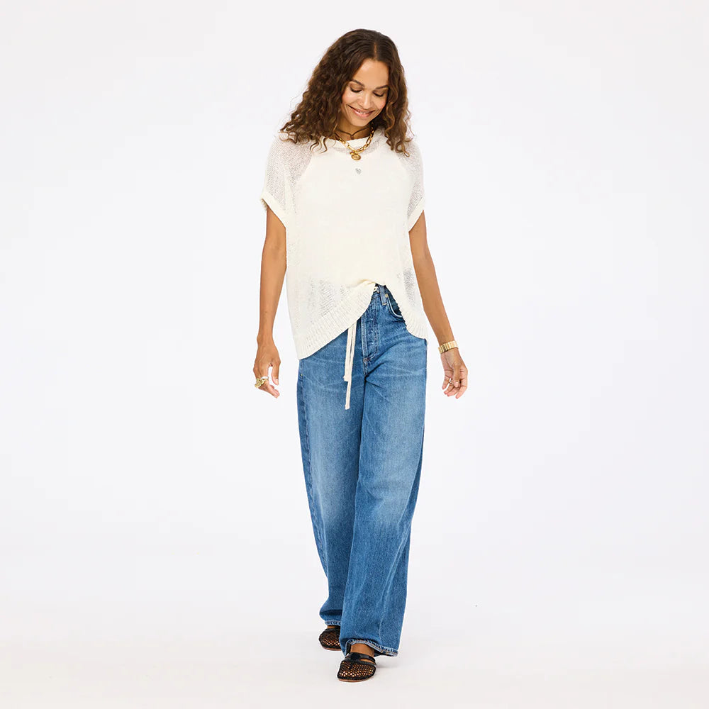 A woman with curly hair smiles and walks against a white background, wearing the Kerri Rosenthal Summer Vestie Sweater, blue wide-leg jeans, and brown shoes.