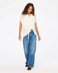 A woman with curly hair smiles and walks against a white background, wearing the Kerri Rosenthal Summer Vestie Sweater, blue wide-leg jeans, and brown shoes.