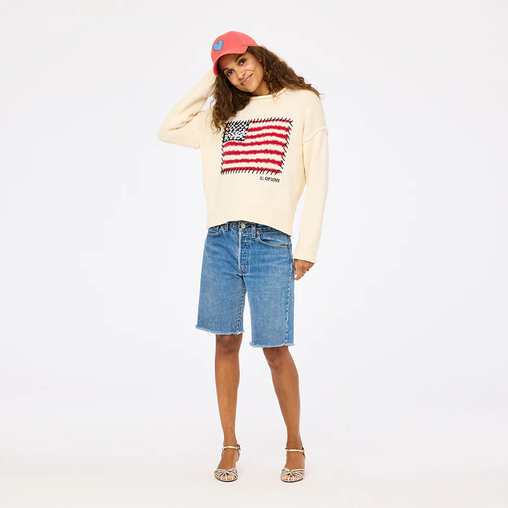 A woman smiles against a white background in the Kerri Rosenthal Roll Neck / US of Love Sweater, denim knee-length shorts, strappy sandals, and a red baseball cap—a perfect patriotic look.