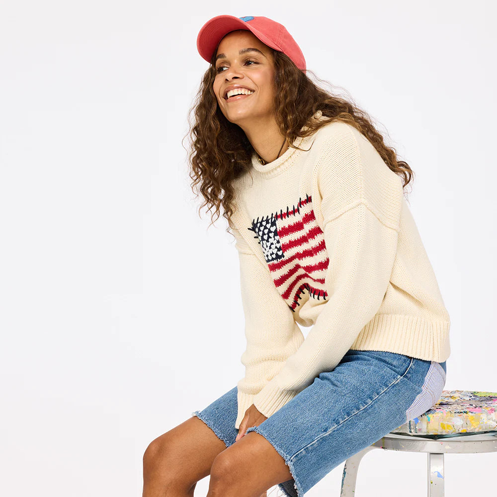 A smiling woman with long curly hair wears the Kerri Rosenthal Roll Neck / US of Love Sweater, denim shorts, and a coral pink cap as she sits on a paint-splattered stool against a plain white background.