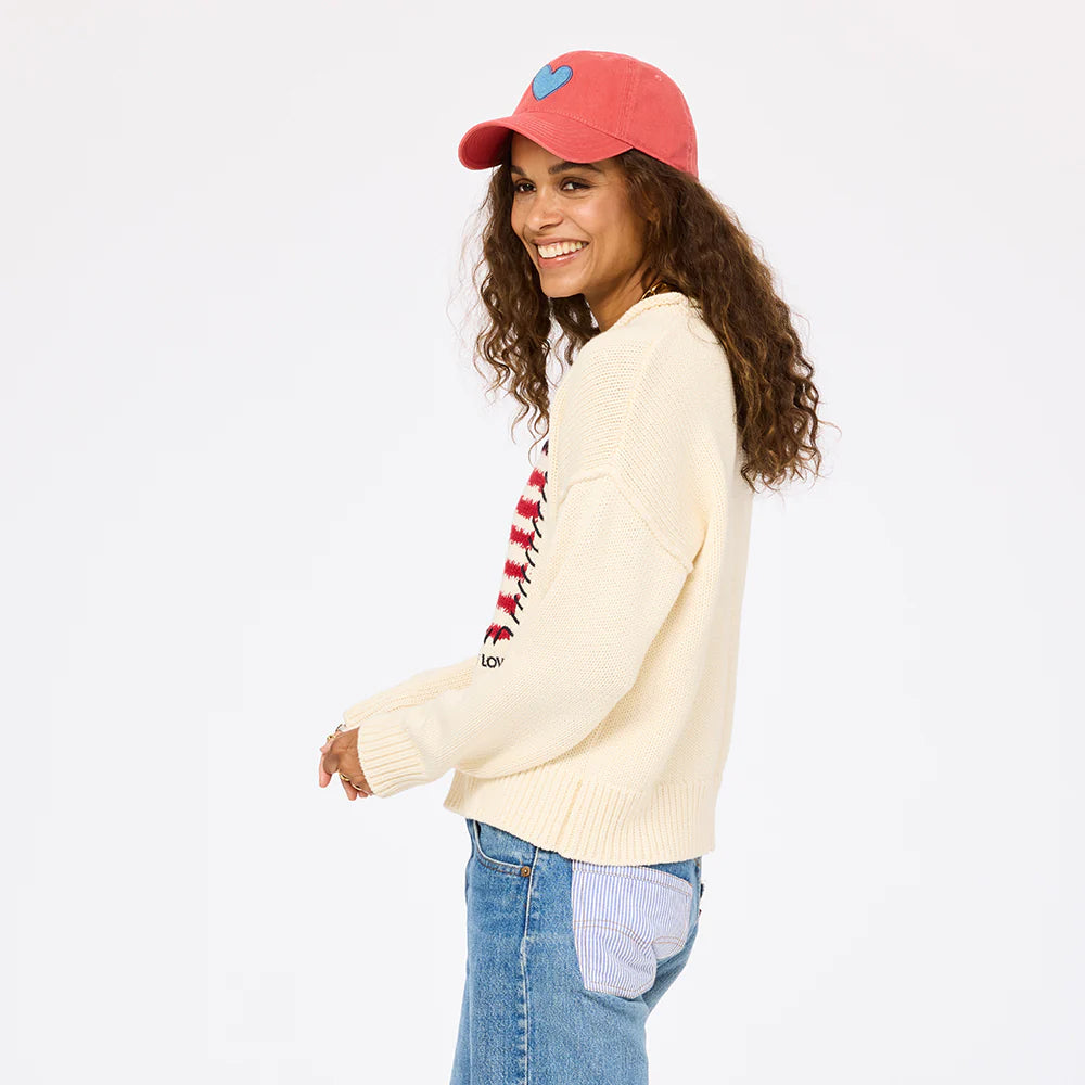 A smiling woman with curly hair wears a pink cap, blue jeans, and the Roll Neck /US of Love Sweater by Kerri Rosenthal. She stands sideways, looking at the camera against a plain white background.