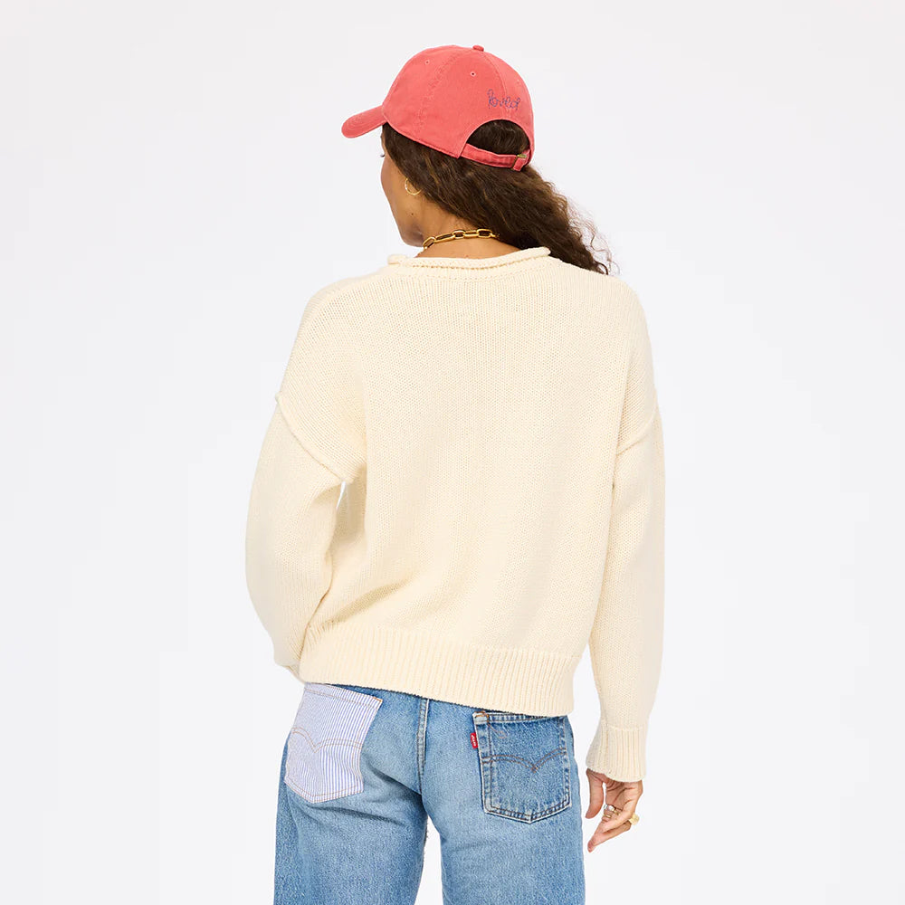 A person with long curly hair wears the Kerri Rosenthal Roll Neck /US of Love Sweater, blue jeans with a striped pocket, and a pink cap while facing away from the camera against a plain white background.