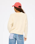 A person with long curly hair wears the Kerri Rosenthal Roll Neck /US of Love Sweater, blue jeans with a striped pocket, and a pink cap while facing away from the camera against a plain white background.