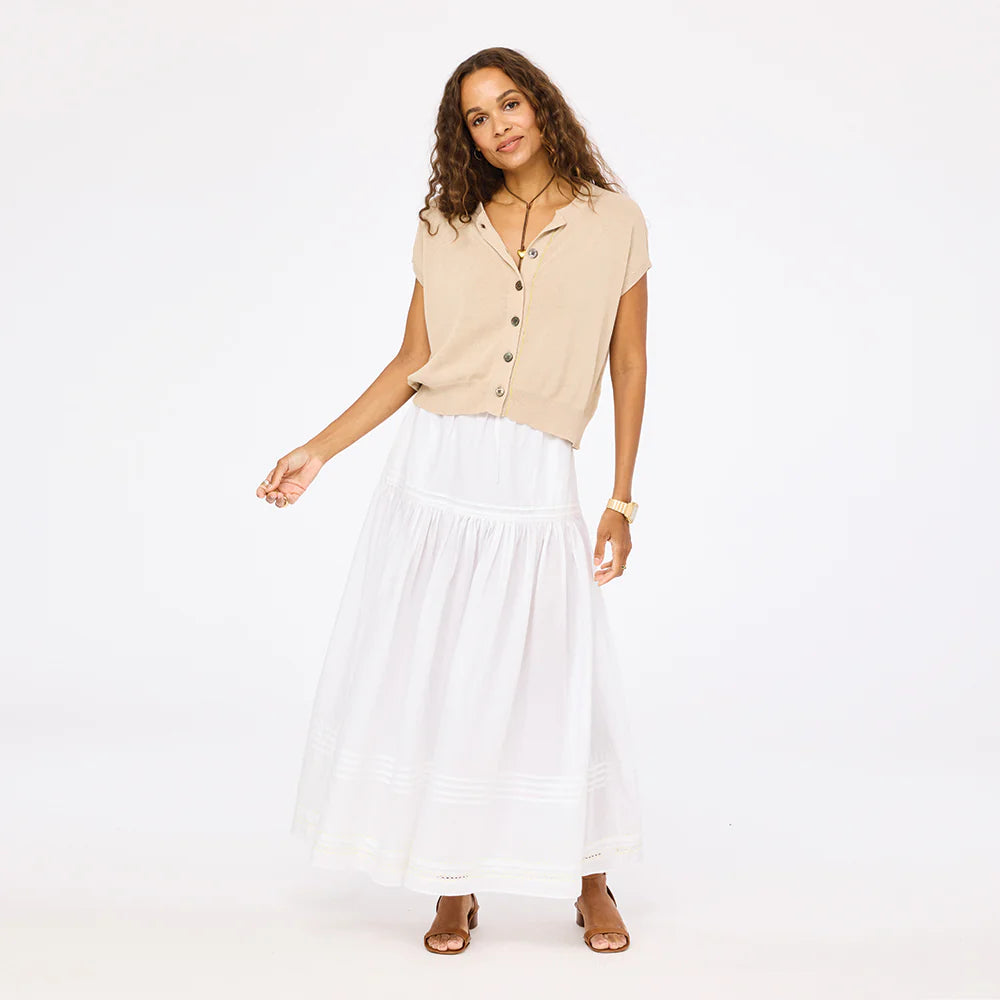 A woman poses against a white background wearing Kerri Rosenthal's Vacance Pintuck Skirt—a long white poplin skirt with tiered lace trim—paired with a beige short-sleeve button-up, brown sandals, and a gold watch for an effortlessly elegant look.