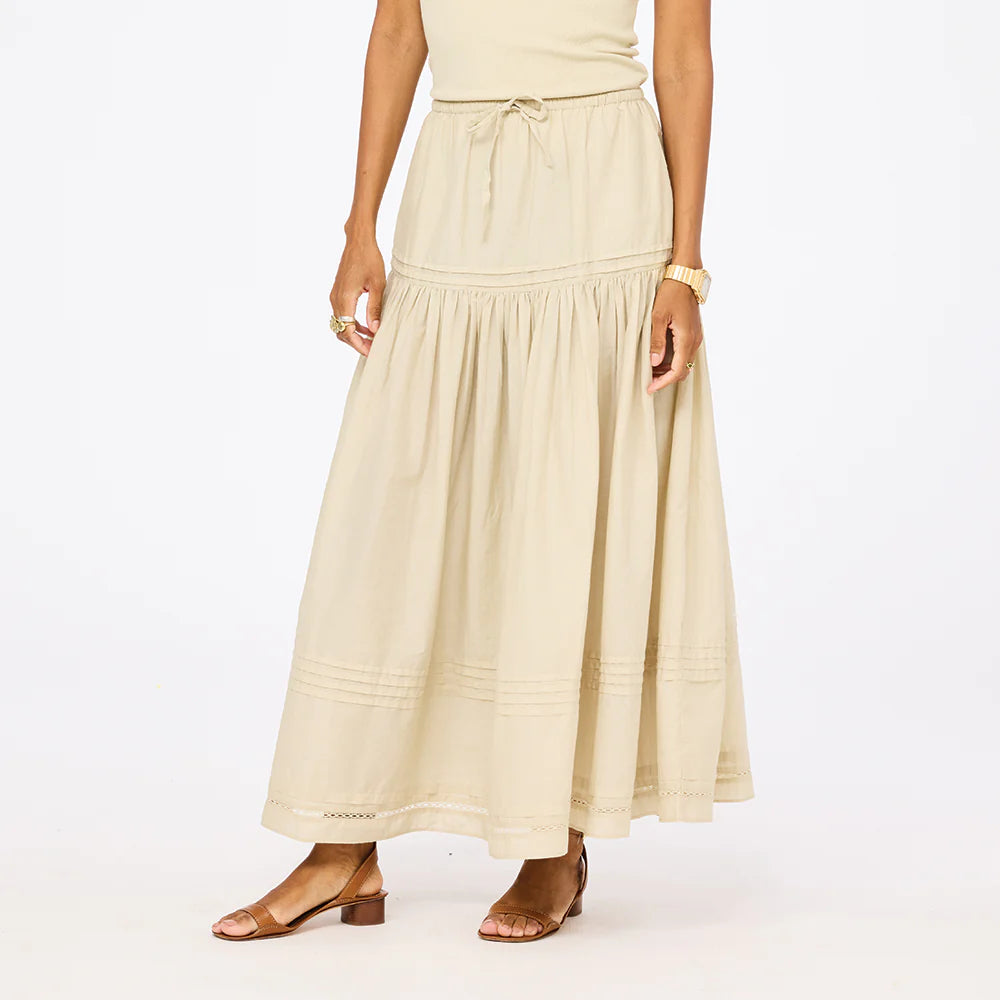 A woman models the Kerri Rosenthal Vacance Pintuck Skirt, featuring tiered lace trim, paired with a sleeveless beige top and brown strappy sandals. She stands against a white background with her face not shown.