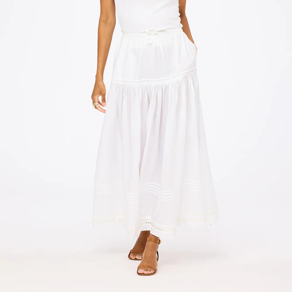 A woman in a sleeveless white top and a Kerri Rosenthal Vacance Pintuck Skirt with sheer details and tiered lace trim, paired with tan heeled sandals, stands against a plain white background.