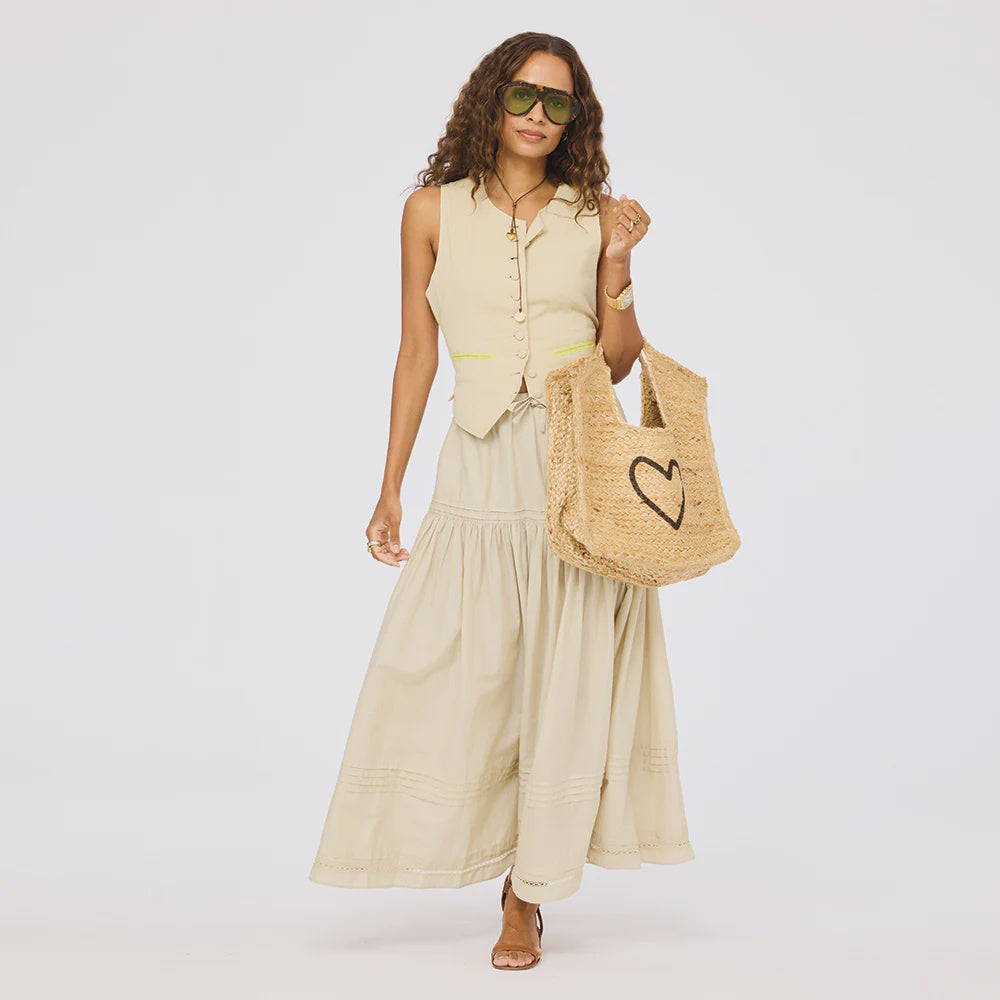 A woman in a sleeveless beige vest, Kerri Rosenthal's Vacance Pintuck Skirt, large sunglasses, and open-toe sandals carries a straw tote bag with a black heart design, standing against a plain white background.