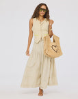 A woman in a sleeveless beige vest, Kerri Rosenthal's Vacance Pintuck Skirt, large sunglasses, and open-toe sandals carries a straw tote bag with a black heart design, standing against a plain white background.