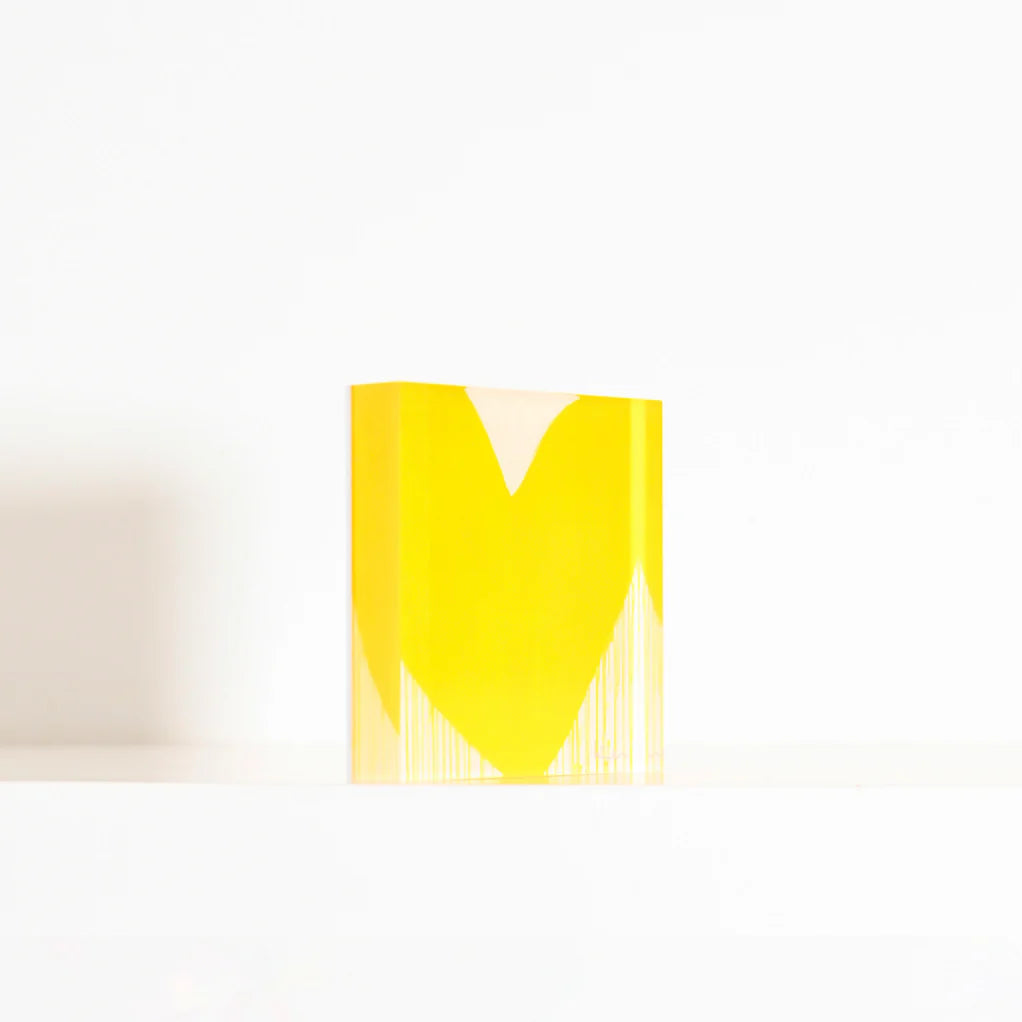 A sculptural piece titled "Mad Love Drippy Heart Block of Love" by Kerri Rosenthal, featuring a heart-like shape at the center, is positioned on a white surface against a plain white background. The yellow geometric design has vertical lines extending downward from the heart shape.