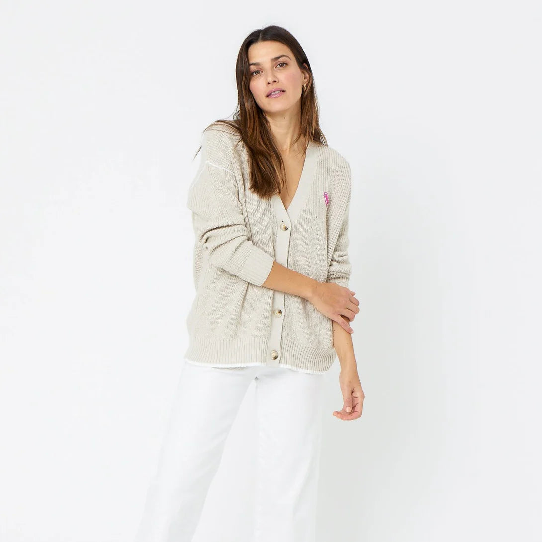 A woman with long, dark hair, wearing a Kerri Rosenthal Slouchy Cardigan in light beige made of 100% cotton, white pants, and featuring a pink heart appliqué on her left side, stands against a plain white background. Her right arm is bent, and her left arm is folded across her body.