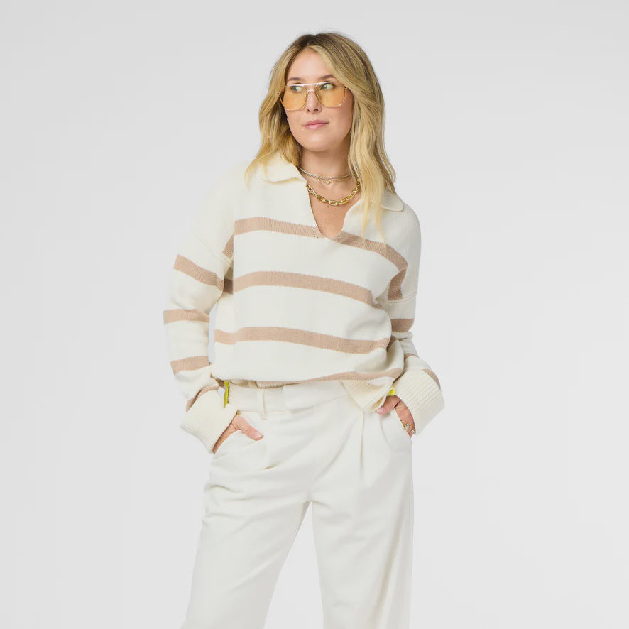 A person with long, blonde hair wears amber-tinted glasses and the Syd Sweater by Kerri Rosenthal, featuring a cream cashmere and cotton design with tan stripes, paired with white pants. They stand with hands in pockets against a plain white background.