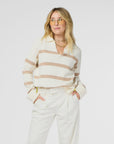 A person with long, blonde hair wears amber-tinted glasses and the Syd Sweater by Kerri Rosenthal, featuring a cream cashmere and cotton design with tan stripes, paired with white pants. They stand with hands in pockets against a plain white background.