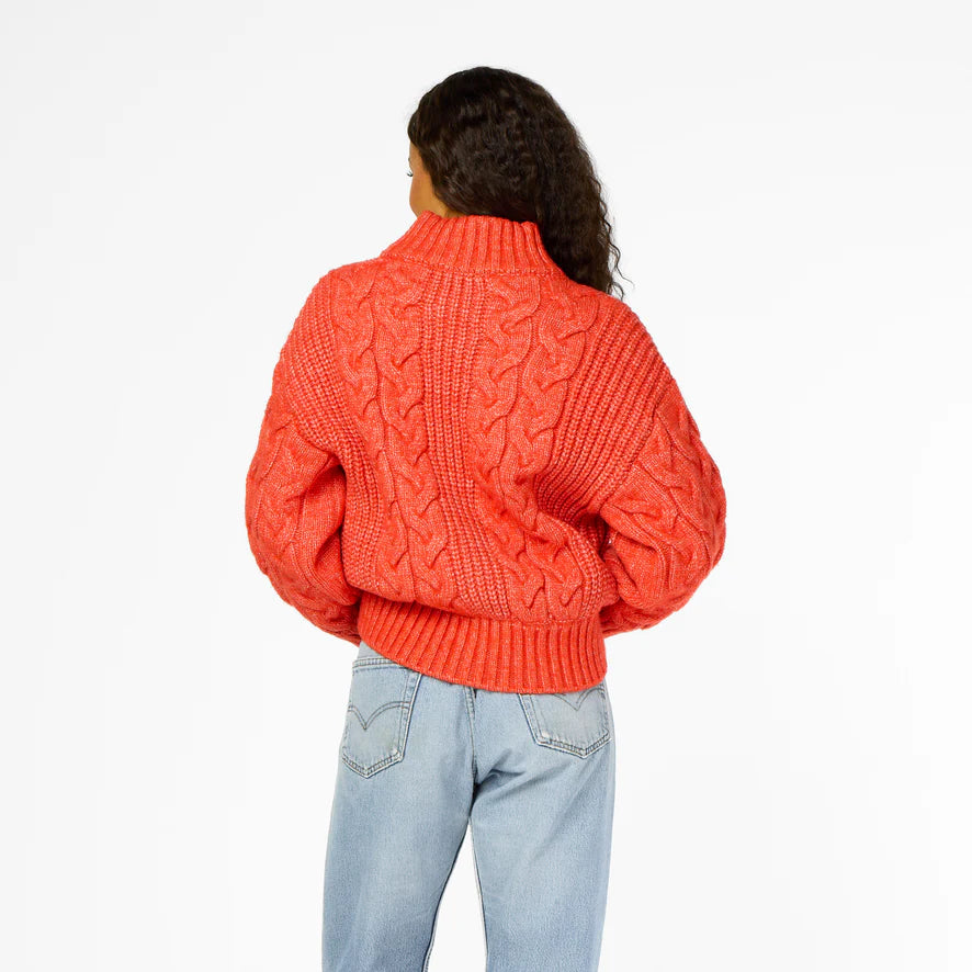 A person with long curly hair, wearing a Kerri Rosenthal Troy Cable Sweater crafted from recycled yarn and light blue jeans, stands facing away. The sweater features a turtleneck and textured pattern against a plain white background.