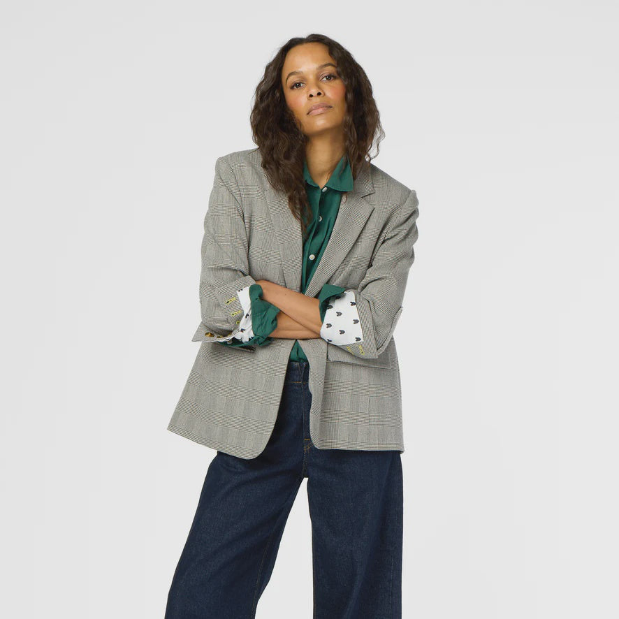 A person confidently stands with arms crossed, clad in a Kerri Rosenthal Workday Plaid Blazer featuring heart patches, over a green shirt with rolled-up sleeves. Their long, wavy hair complements their dark wide-leg jeans against the plain white background as they gaze directly at the camera.