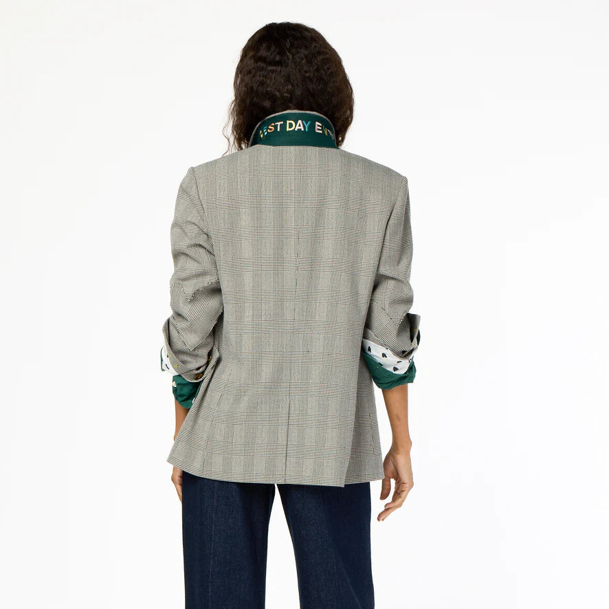 A person with long hair stands facing away from the camera, sporting a Kerri Rosenthal Workday Plaid Blazer over a green shirt with rolled-up sleeves and dark jeans. Some text is visible on the blazer collar, though parts of it are obscured.