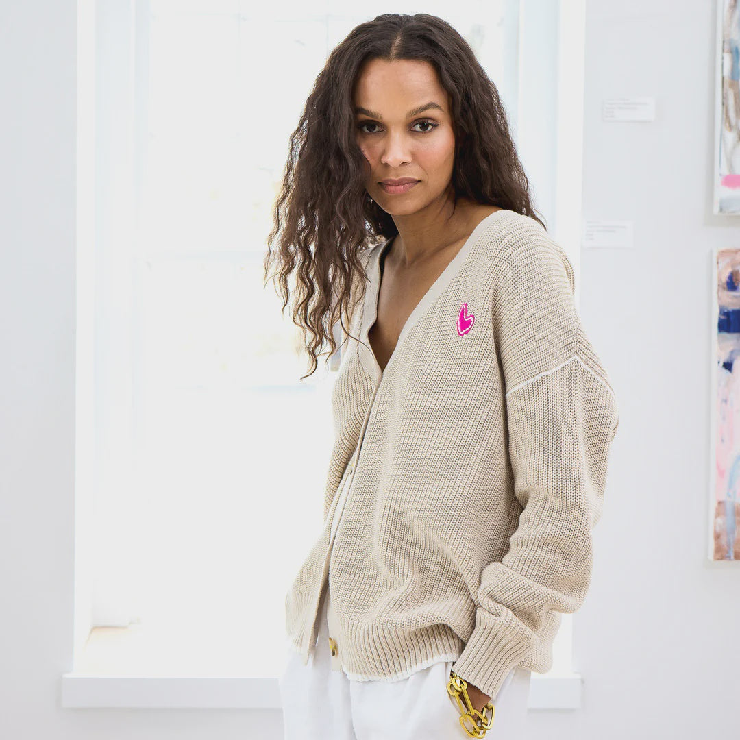 A person with long wavy hair is wearing a Kerri Rosenthal Slouchy Cardigan with a pink heart appliqué on the chest, and white trousers. They are standing indoors in a brightly lit room, looking towards the camera with a neutral expression.