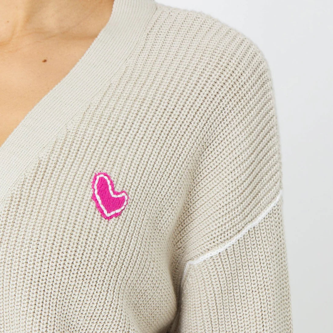 Close-up of a person wearing the Kerri Rosenthal Slouchy Cardigan in light beige, featuring a pink heart-shaped appliqué on the left side. The focus is on the upper left part of this 100% cotton cardigan, highlighting its texture and the embroidered heart design.