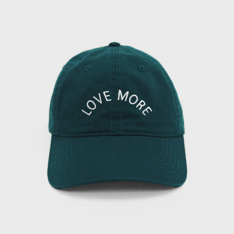 A dark emerald green Love More Hat by Kerri Rosenthal, crafted from 100% cotton, features white embroidered text on the front that reads "LOVE MORE." This baseball hat includes a curved brim and visible stitching lines. The background is plain white.