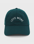 A dark emerald green Love More Hat by Kerri Rosenthal, crafted from 100% cotton, features white embroidered text on the front that reads "LOVE MORE." This baseball hat includes a curved brim and visible stitching lines. The background is plain white.