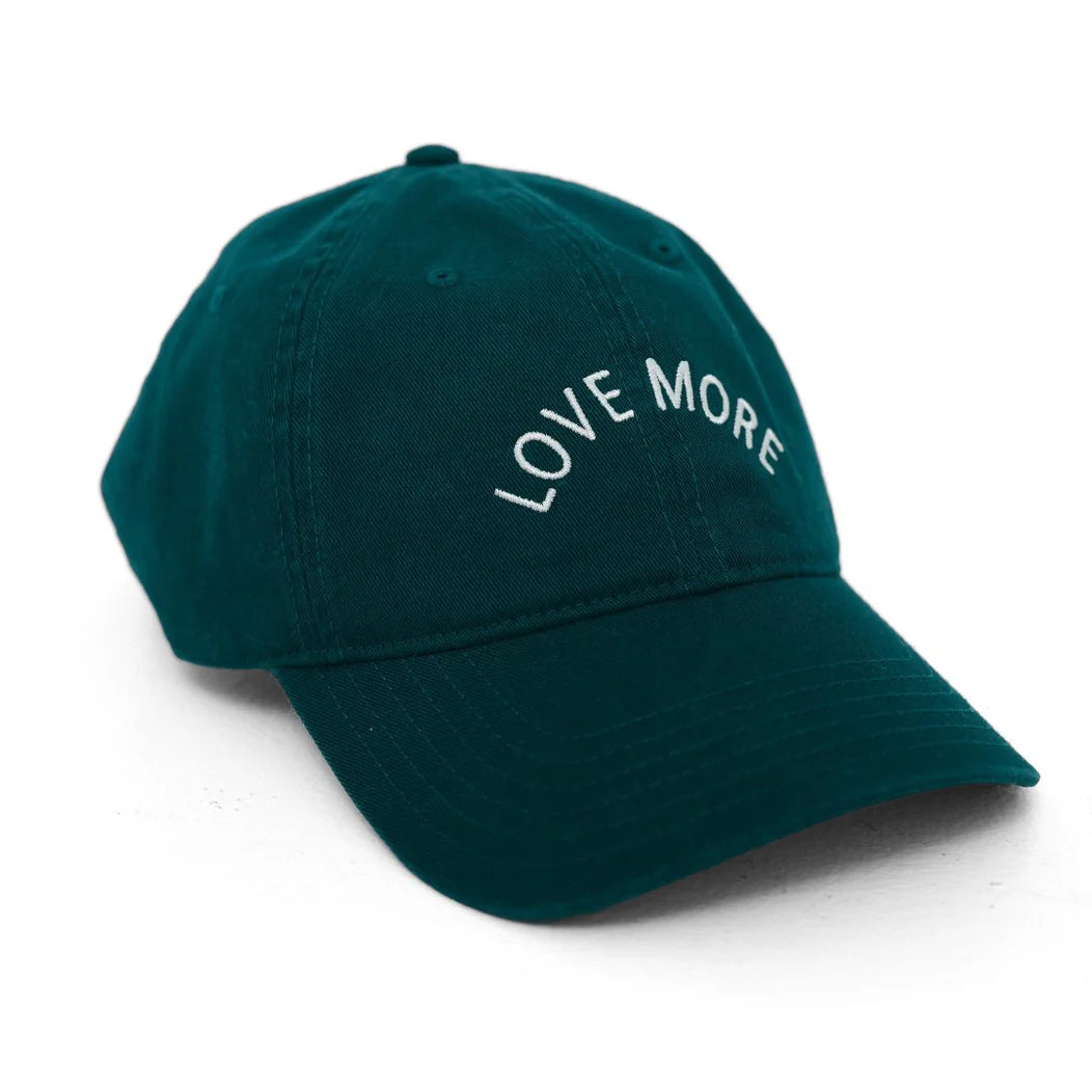 A Kerri Rosenthal Love More Hat, featuring an emerald green color and made from 100% cotton, showcases the phrase "LOVE MORE" embroidered in white text on the front panel. This baseball cap is presented on a white background.