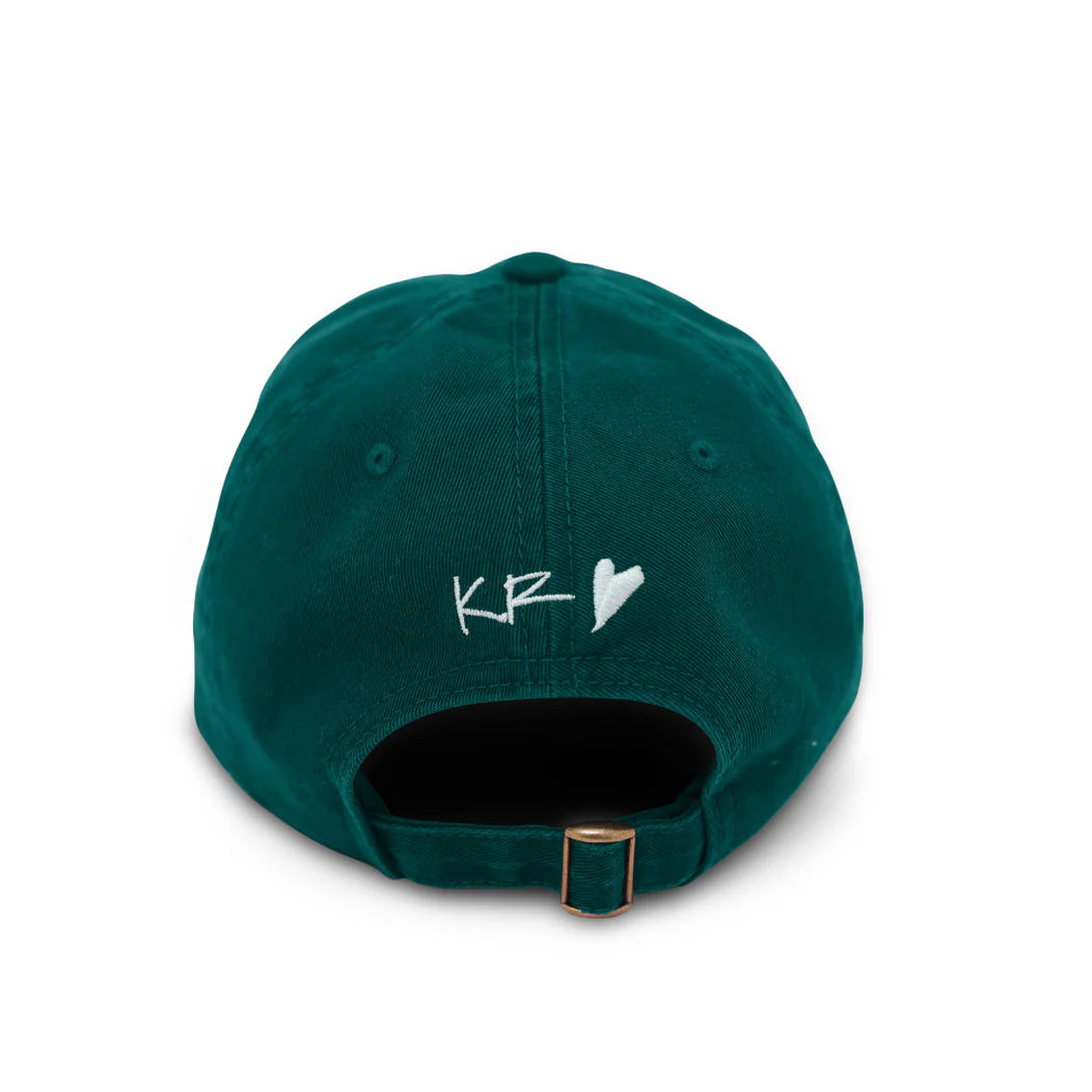 A dark green baseball cap, known as the Love More Hat by Kerri Rosenthal, is shown from the back. This 100% cotton cap features a metal buckle closure and white embroidered letters "KR" followed by a heart on the fabric just above the opening.