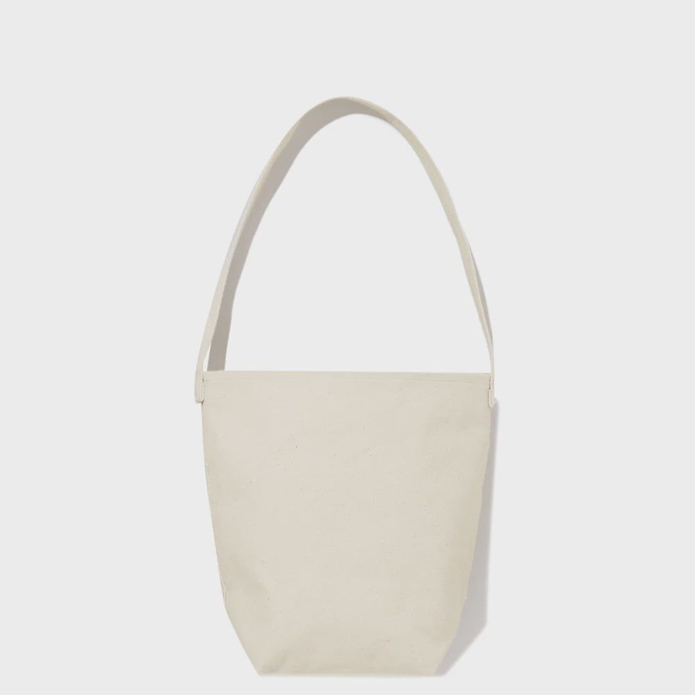 The Uma by Kule is a minimalist beige canvas bucket bag made from 100% cotton, featuring a single shoulder strap. It's showcased against a plain white background with a simple design and no visible pockets or embellishments.