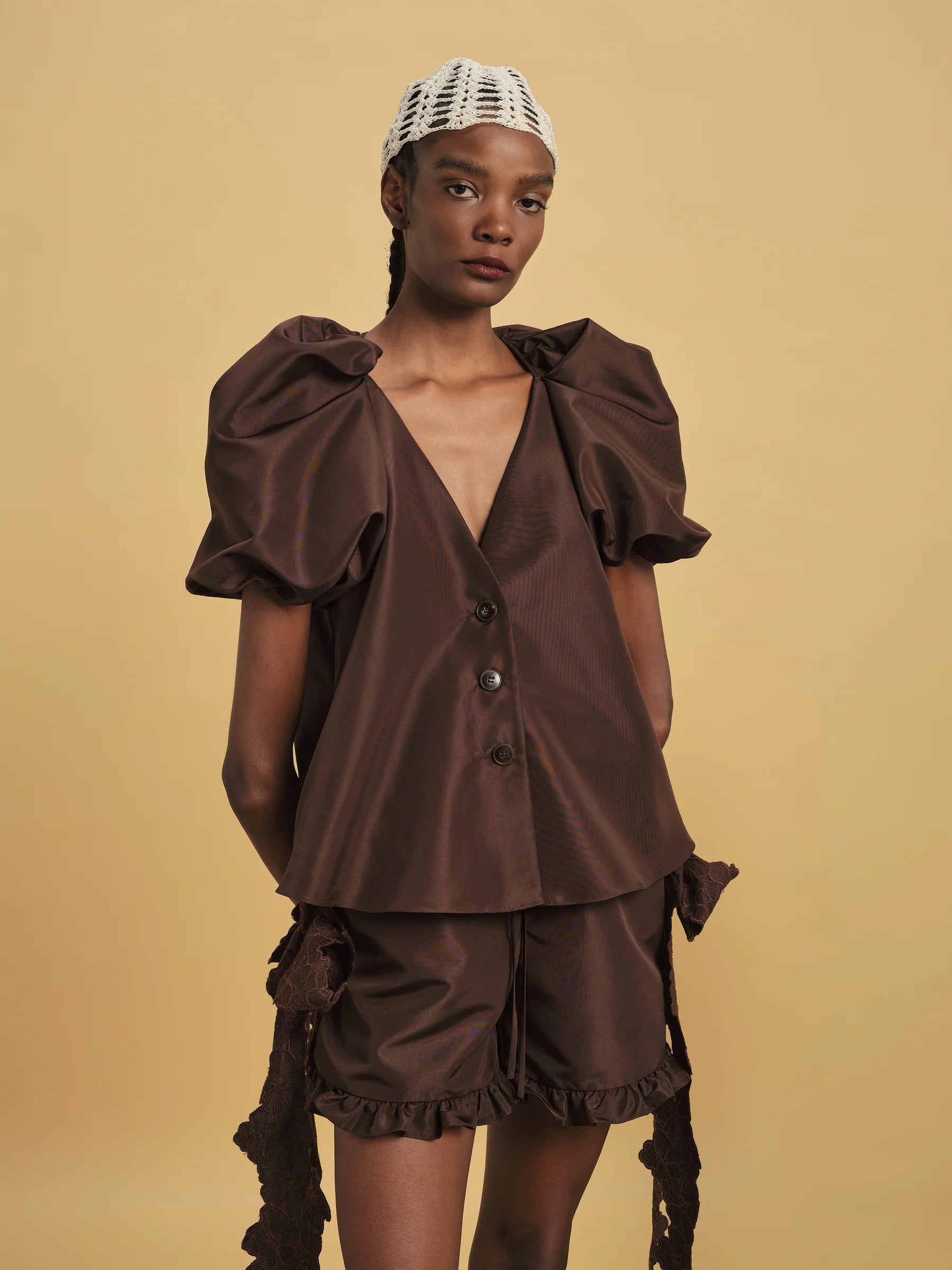 Against a beige background, someone wears the Kika Vargas Harpa Top in brown—featuring voluminous sleeves and a deep V-neck—styled with matching brown shorts and a white crocheted headpiece.