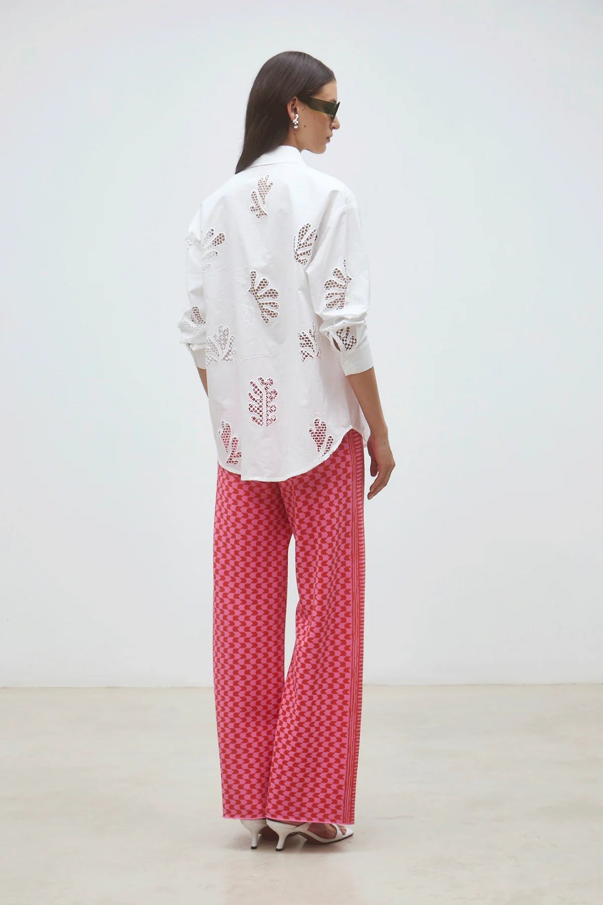 A woman with long dark hair, sunglasses, and white heels stands in a minimal room wearing the SUNCOO Loula Blouse—an embroidered white 100% organic cotton button-down—and loose pink checkered pants. She is facing away.