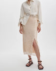 A person wearing a cream button-up shirt with rolled sleeves, the kali skirt by Rag & Bone—a light beige sequin midi skirt with a front slit—and brown strappy flat sandals, standing against a plain white background.
