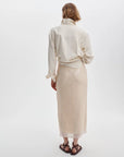 A person with short, wavy hair faces away, styled in a cream jacket and Rag & Bone's shimmering kali skirt, paired with black sandals against a plain white background.