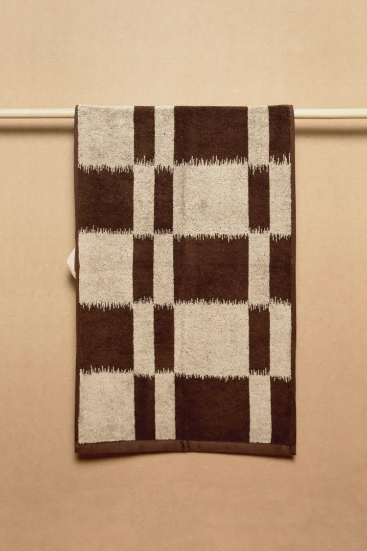 The Karin Hand Towel by Autumn Sonata, made from organic cotton and featuring a bold brown and beige plaid pattern inspired by Ikat weaving, hangs neatly on a rod against a beige background.