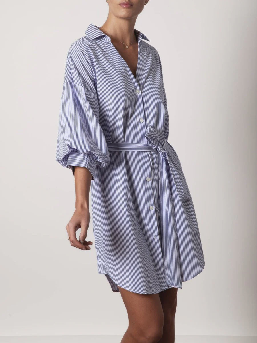 A woman wears the KATE BELTED DRESS by BROCHU WALKER, a loose-fitting blue and white vertical striped shirtdress with long sleeves and a tied waist belt, standing against a plain light background.