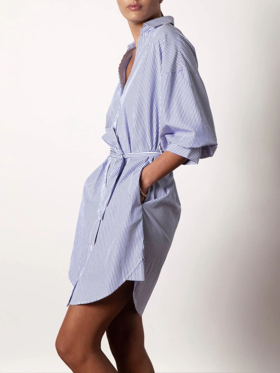 A person wearing the BROCHU WALKER KATE BELTED DRESS, featuring blue and white vertical stripes, a belt, and rolled-up sleeves, stands sideways with one hand in the pocket against a plain background.