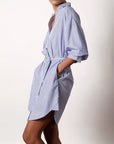 A person wearing the BROCHU WALKER KATE BELTED DRESS, featuring blue and white vertical stripes, a belt, and rolled-up sleeves, stands sideways with one hand in the pocket against a plain background.