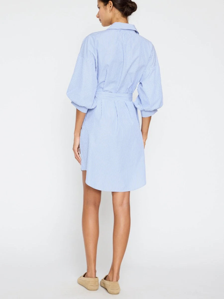 A woman with her hair tied up models the BROCHU WALKER KATE BELTED DRESS, a light blue shirtdress with blouson sleeves and white pinstripes, paired with beige slip-on shoes against a plain white background.