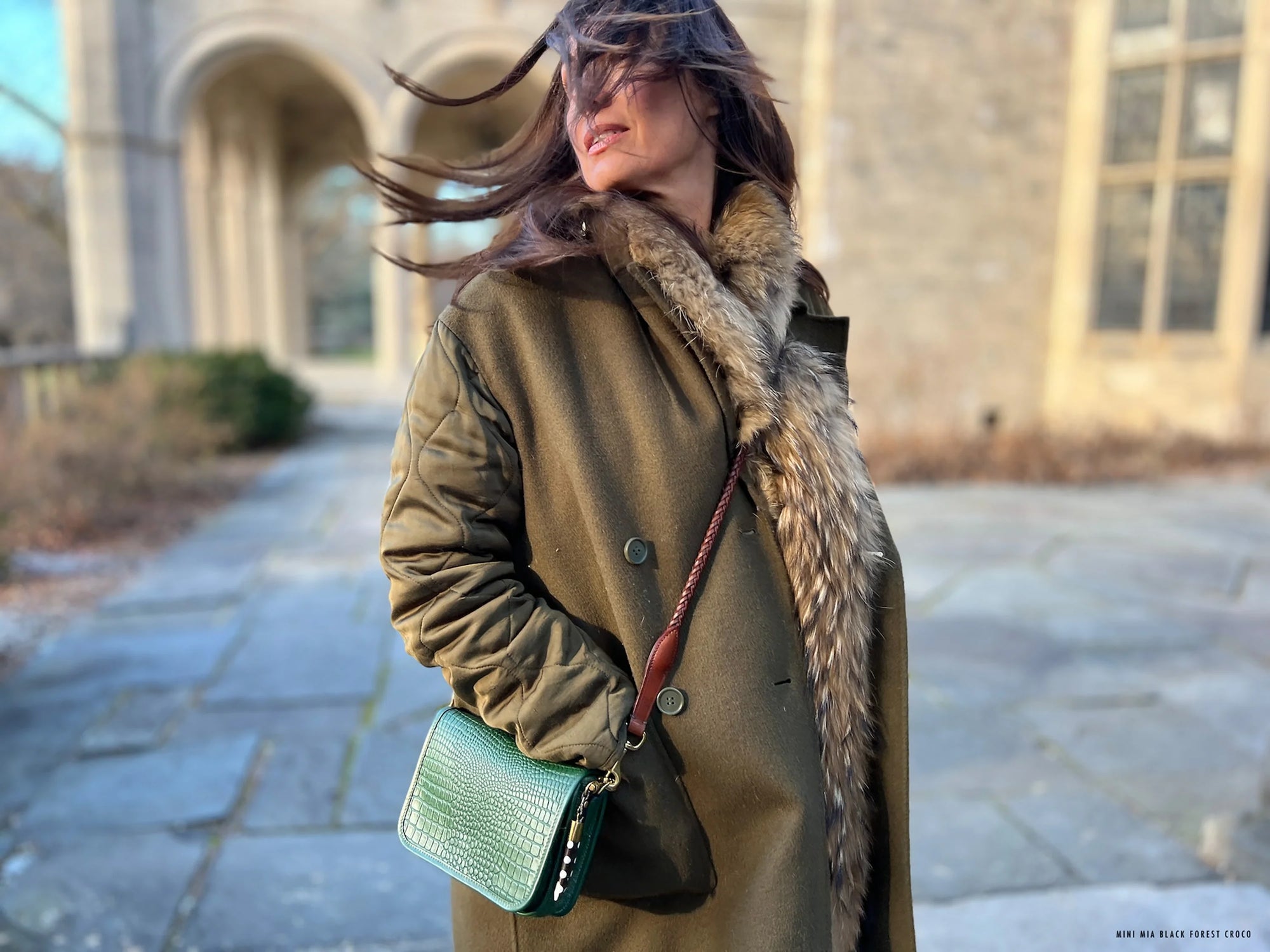 A woman in a green coat with a fur collar stands on a stone path, hair blowing in the wind, holding the Kempton & Co. Mini Mia Crossbody clutch. An old stone building appears in the background.