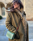A woman in a green coat with a fur collar stands on a stone path, hair blowing in the wind, holding the Kempton & Co. Mini Mia Crossbody clutch. An old stone building appears in the background.