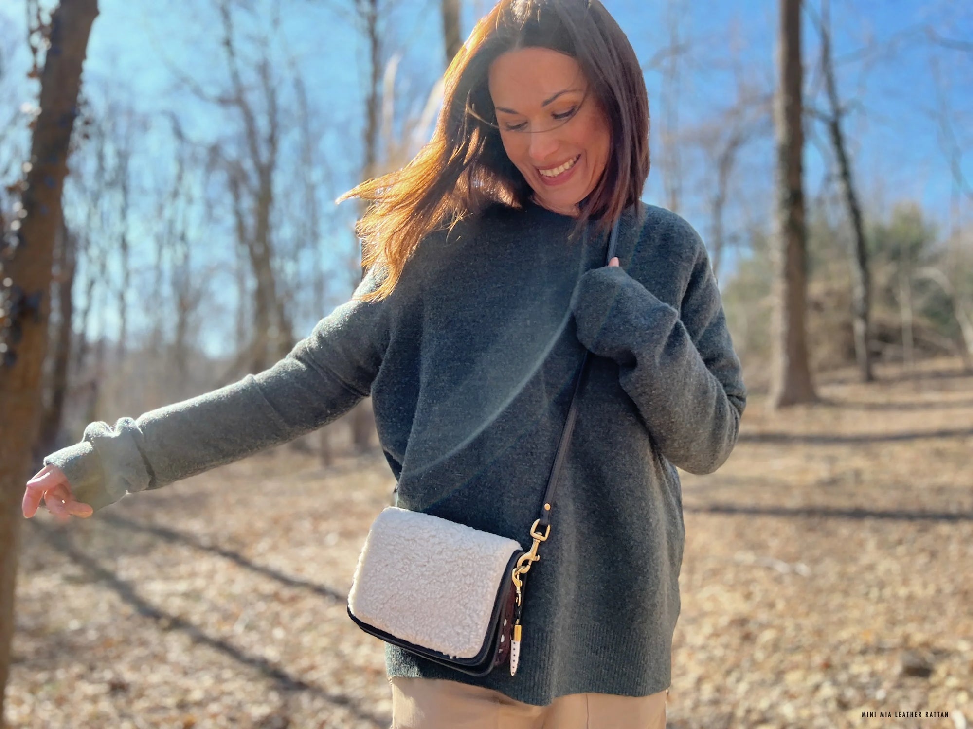 A woman in a gray sweater and light pants smiles in a sunlit, leafless forest, carrying the Mini Mia Crossbody by Kempton &amp; Co. as she enjoys the outdoors on a bright day.