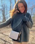 A woman in a gray sweater and light pants smiles in a sunlit, leafless forest, carrying the Mini Mia Crossbody by Kempton & Co. as she enjoys the outdoors on a bright day.