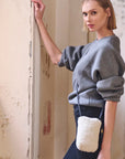 A person with short blonde hair leans against a wall, wearing a gray sweater and black pants. They carry a small Mini Sling Bag Natural Shearling from Kempton & Co., featuring a sleek leather strap. The setting appears to be an indoor space with peeling paint on the walls.