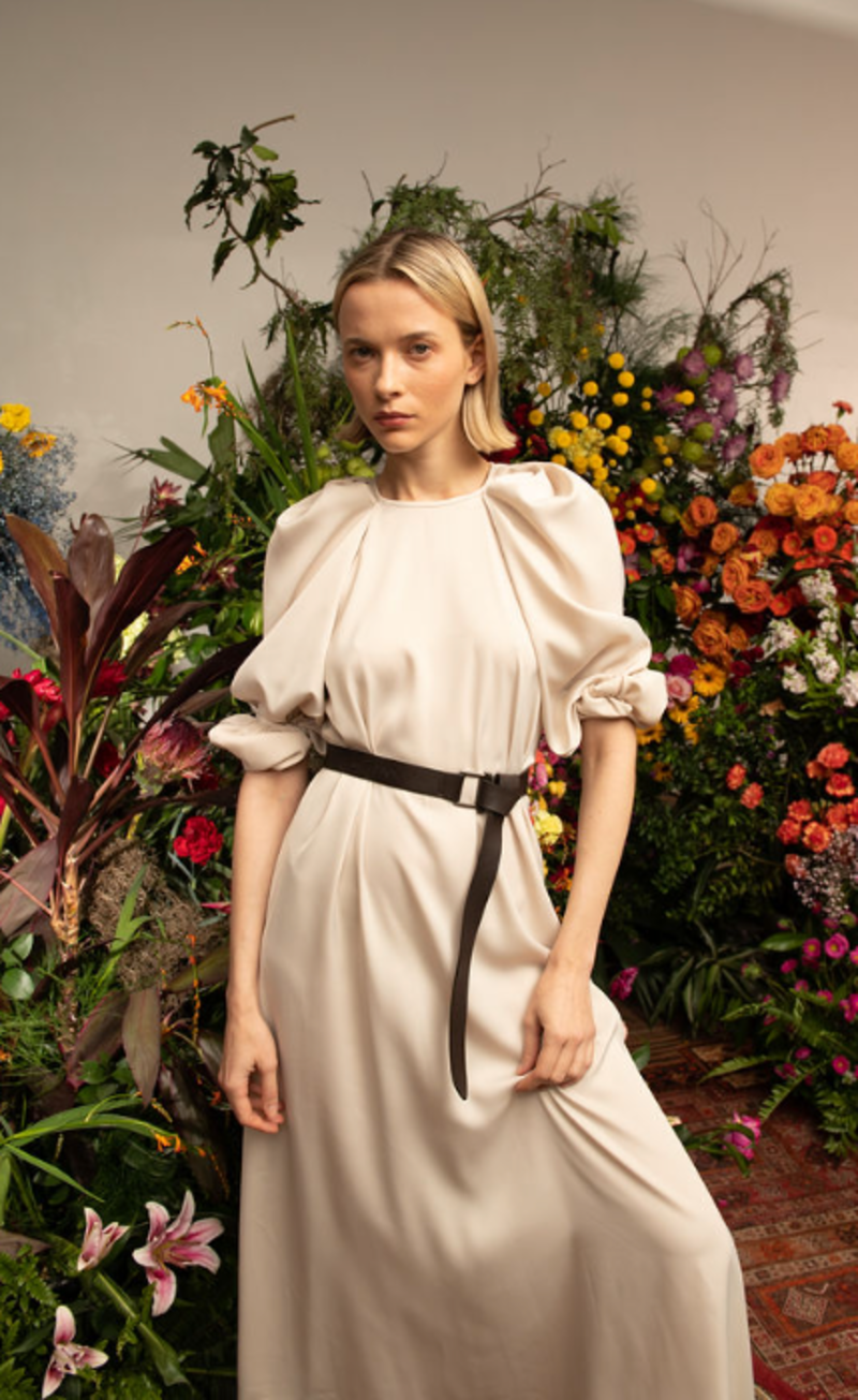 A woman in a cream satin Xeni Dress by Kika Vargas, detailed with puffed sleeves and a dark belt, stands before a vibrant floral display.