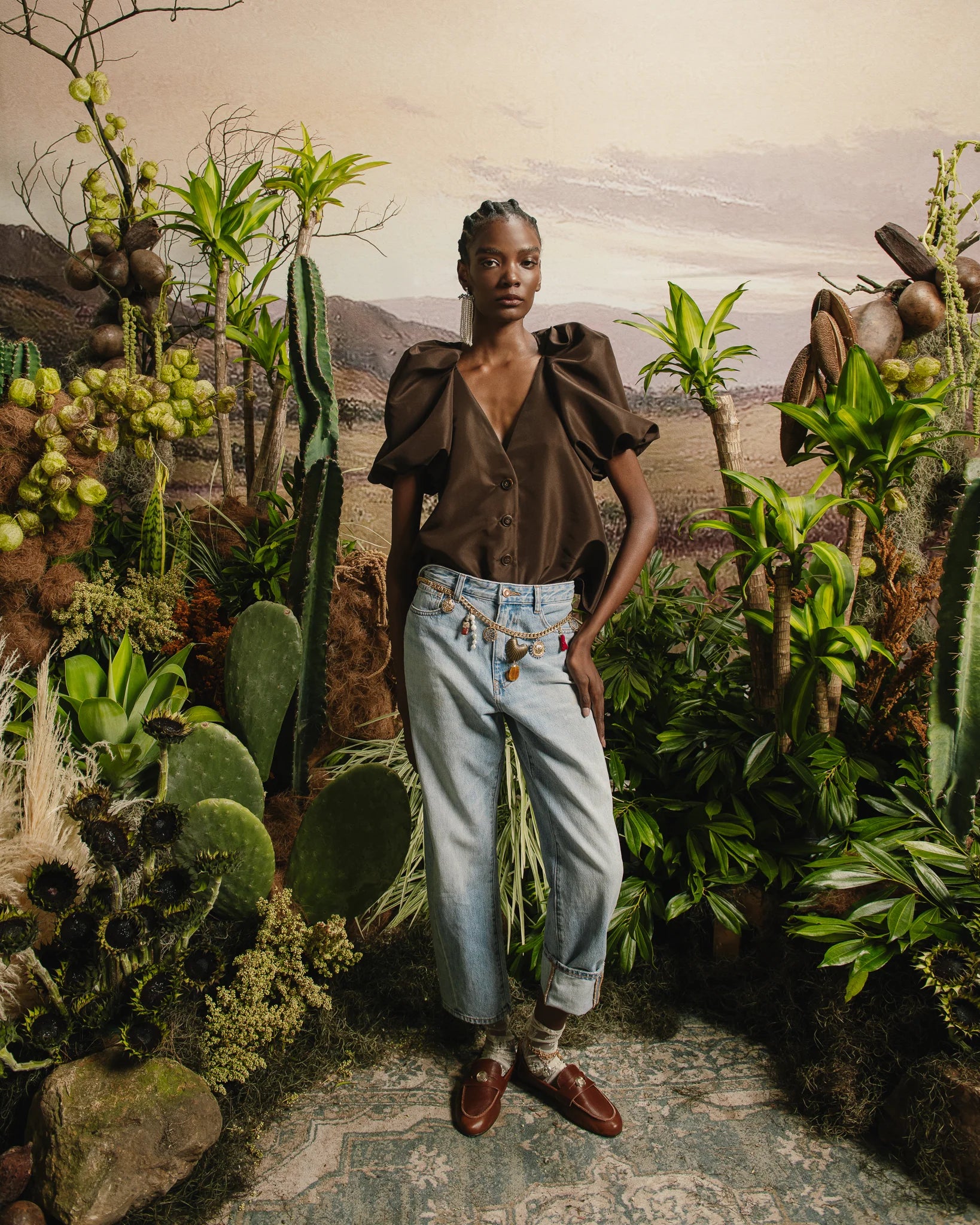 Amid lush greenery and cacti, a person models the Kika Vargas Harpa Top with voluminous sleeves, paired with light blue cuffed jeans, brown loafers, and a decorative belt against a desert backdrop.