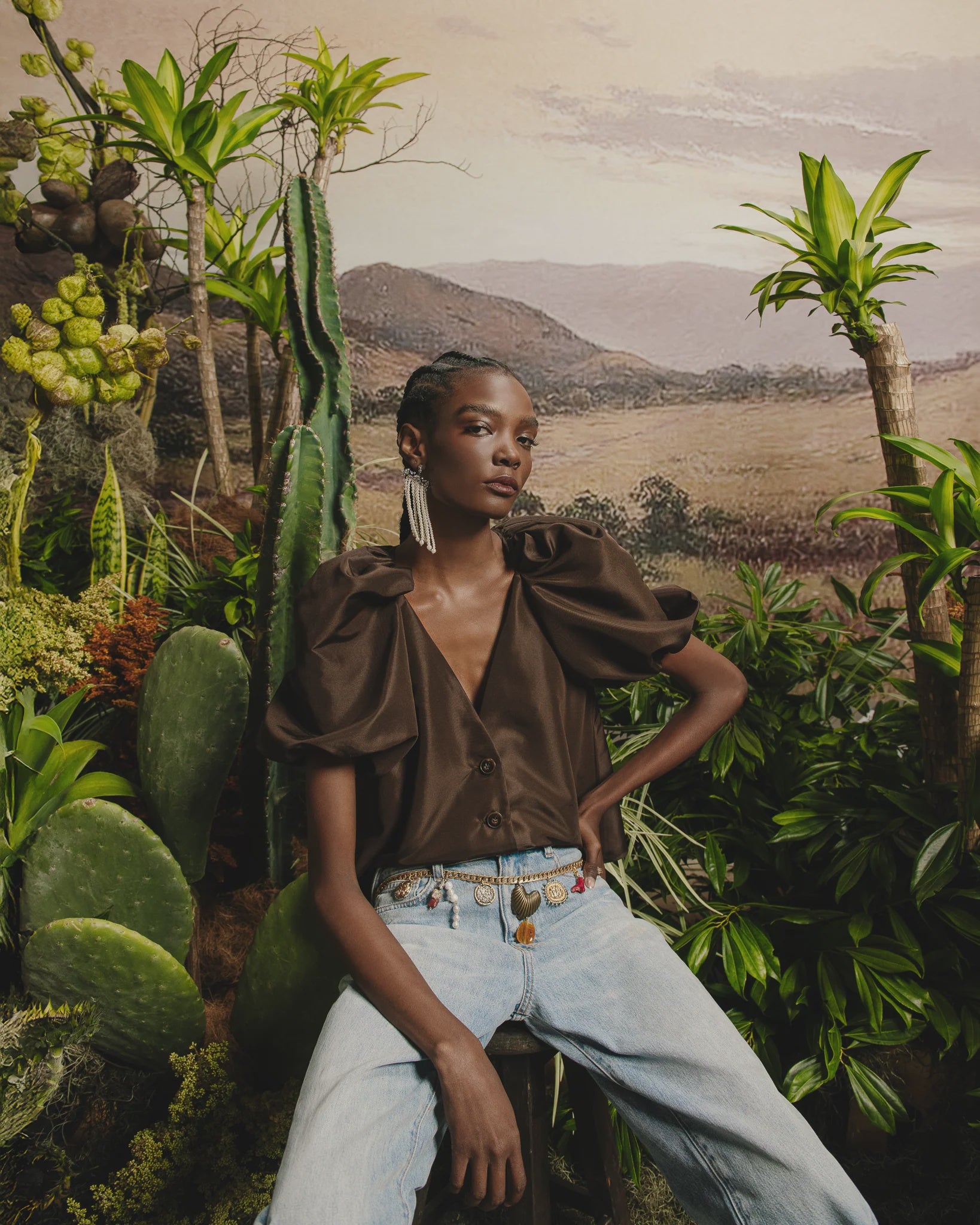 Wearing a dark brown Kika Vargas Harpa Top with voluminous sleeves, light blue jeans, and statement earrings, a person poses among green plants and cacti, with scenic hills in the background.