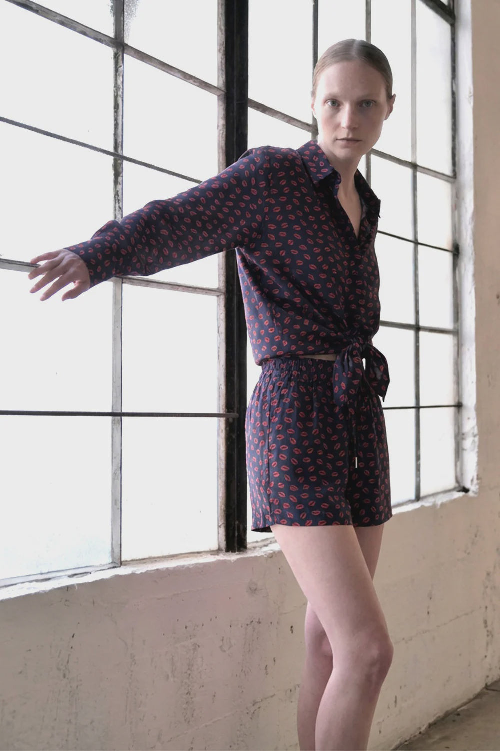A person stands indoors by large windows, wearing Le Superbe's Many Kisses Boxer and a matching dark button-up shirt with a red kiss print, one arm extended, looking at the camera.