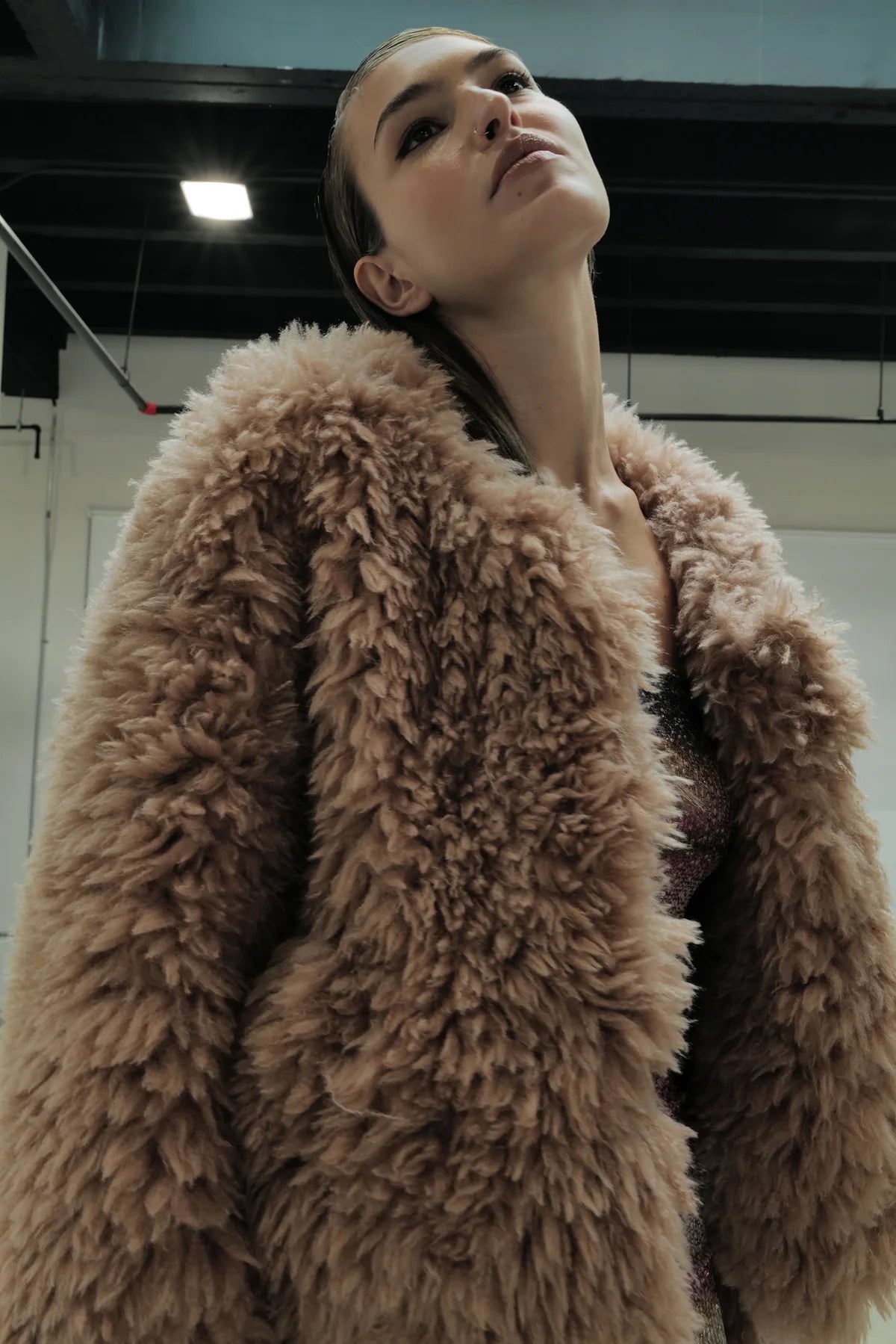 A person wearing the Madison Chubby jacket by Le Superbe in light brown plush faux shearling looks upward in a modern indoor space with exposed ceilings and bright lighting.